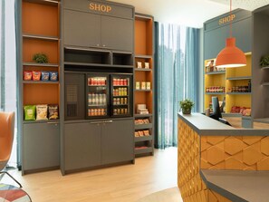 Una tienda con estantes llenos de aperitivos y bebidas, una vitrina refrigerada y una zona de recepción con mostrador de madera.