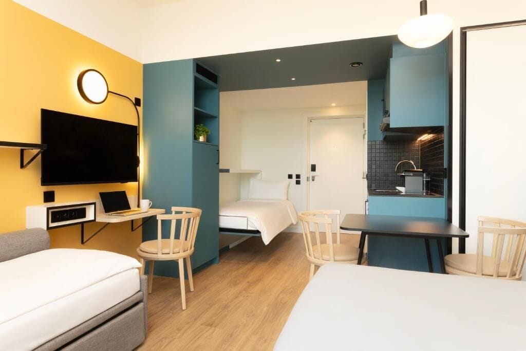 A compact hotel room with a bed, a desk with a laptop, a TV, and a kitchenette.