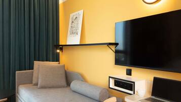 A modern room with a grey sofa, a wooden chair, a desk with a laptop, a TV mounted on the wall, and a framed picture above the TV.