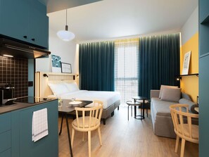 Habitación de hotel con cama, una pequeña mesa de comedor, sofá, zona de cocina y ventana con cortinas.