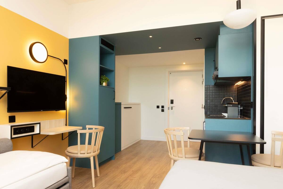 A modern hotel room with a flat-screen TV, a dining table, two chairs, a sofa, and a kitchenette.