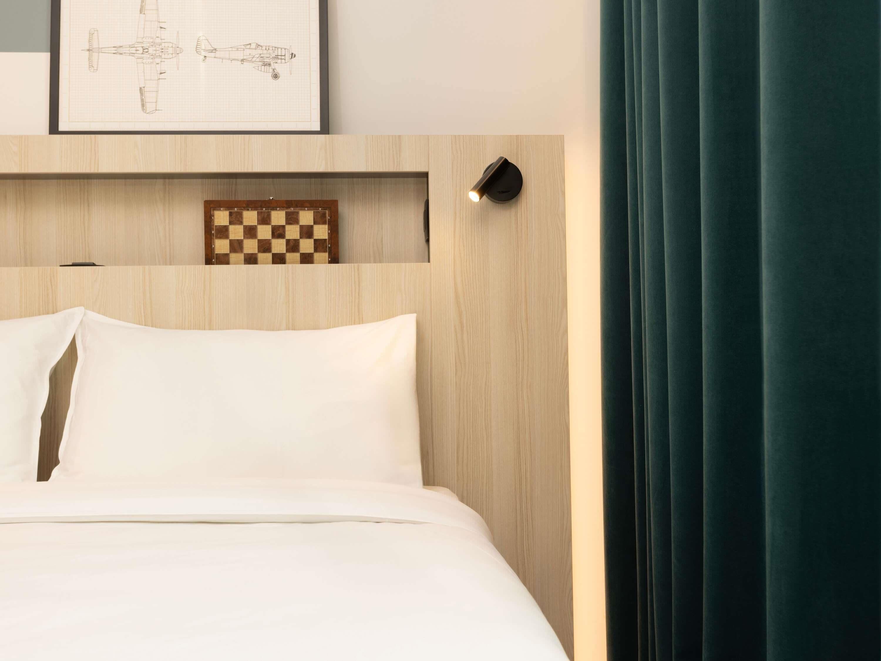 A neatly made bed with white linens, a wooden headboard, a framed chessboard, and a wall-mounted light fixture.