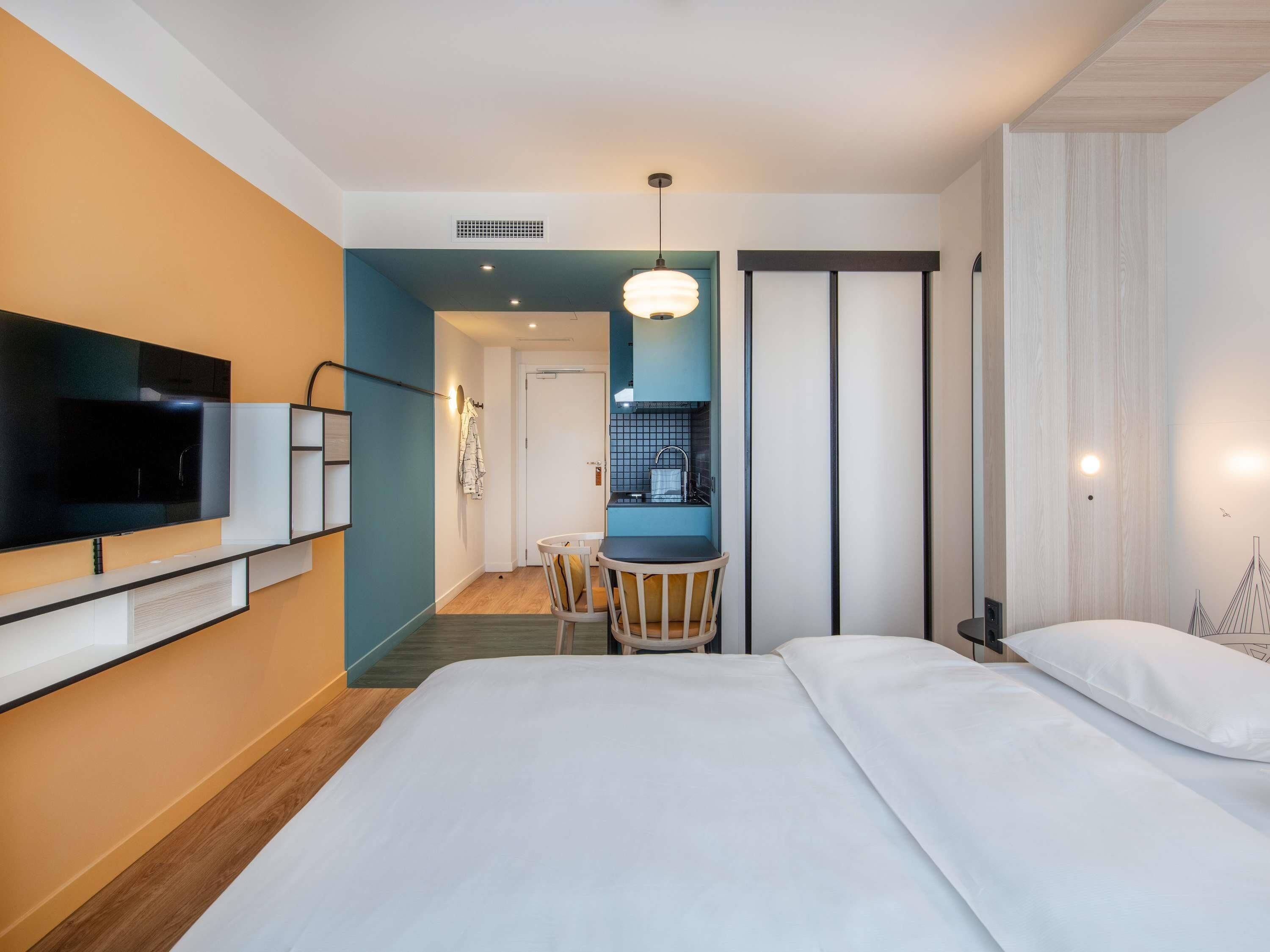 Ein Hotelzimmer mit einem Bett, einem an der Wand befestigten Fernseher, einem Schreibtisch mit Stuhl und einer Küchenzeile.