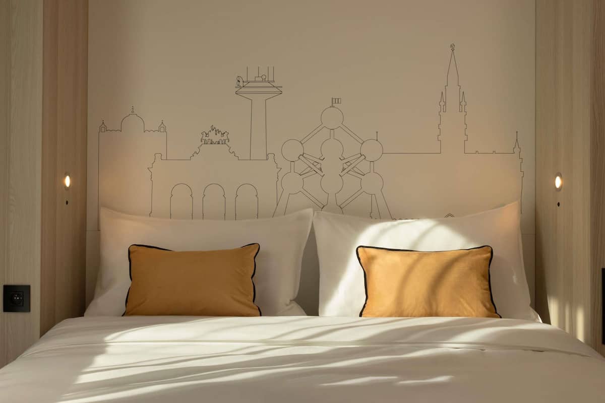 A bed with white bedding and two gold pillows, a wall with a cityscape mural, and a light switch on the wall.