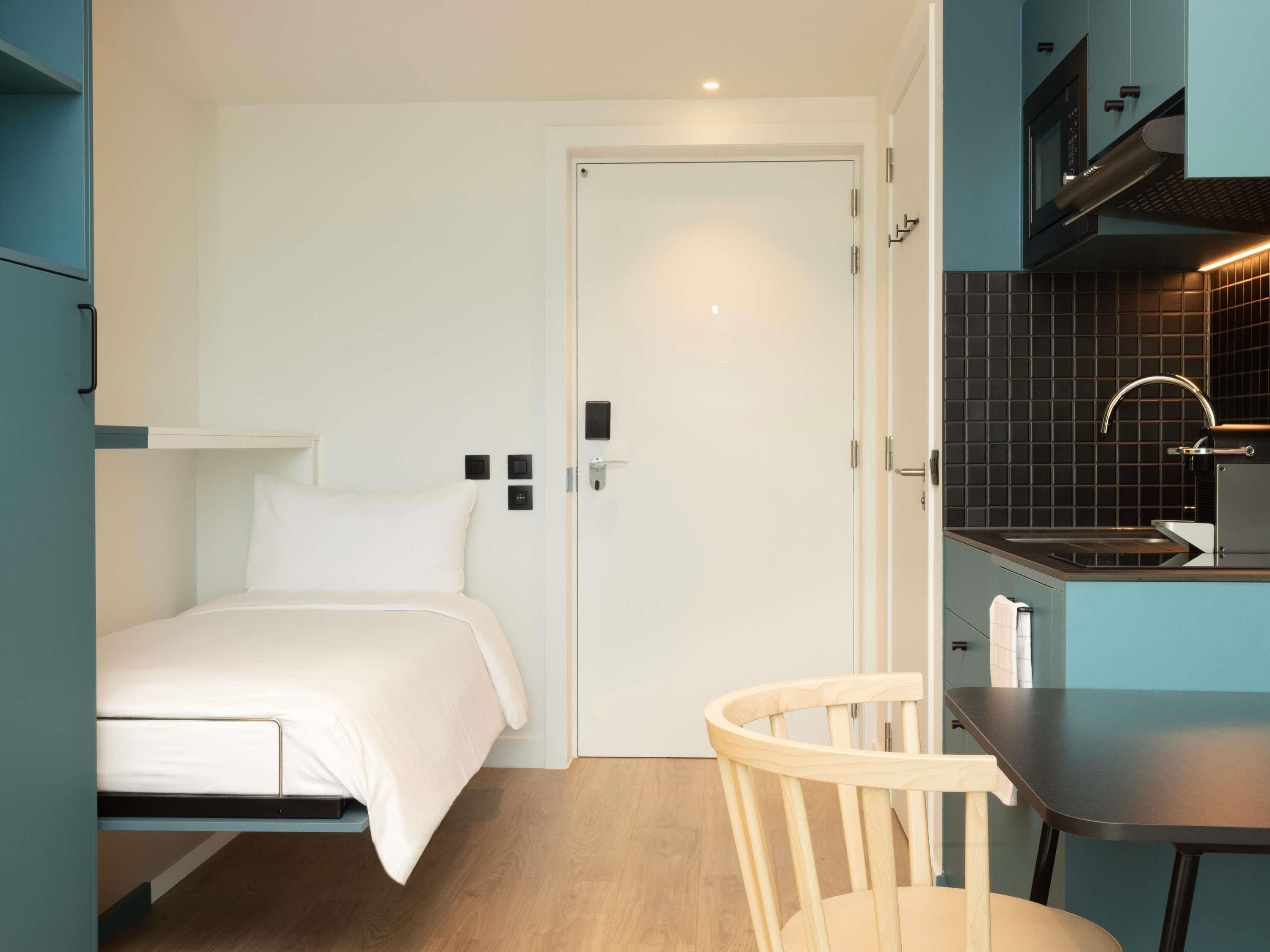 A compact room with a bed, a small table, a chair, a kitchenette with a sink and stove, and a door.