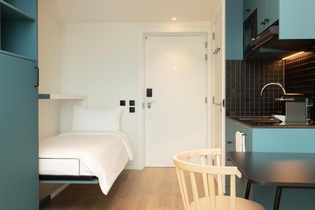 Una habitación compacta con cama, una mesita, una silla y un kitchenette.