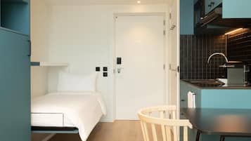 Una habitación compacta con cama, una mesita, una silla y un kitchenette.