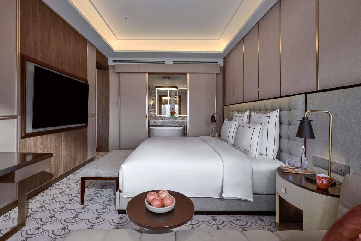 Une chambre d’hôtel moderne dotée d’un grand lit, d’un téléviseur à écran plat, d’une table de chevet avec une lampe et d’une petite table ronde sur laquelle sont posées des pommes.