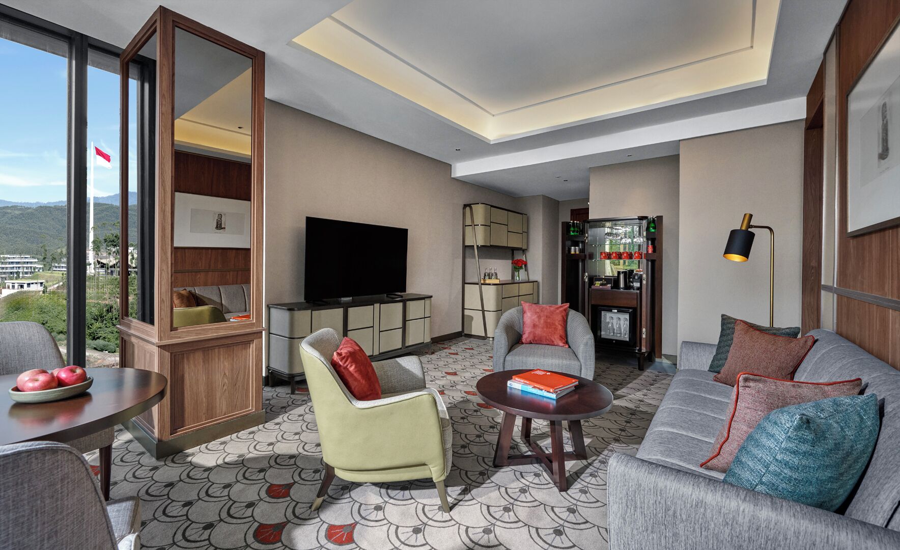 A modern hotel room with a large window, a flat-screen TV, a sofa, a chair, a small table, and a bookshelf.