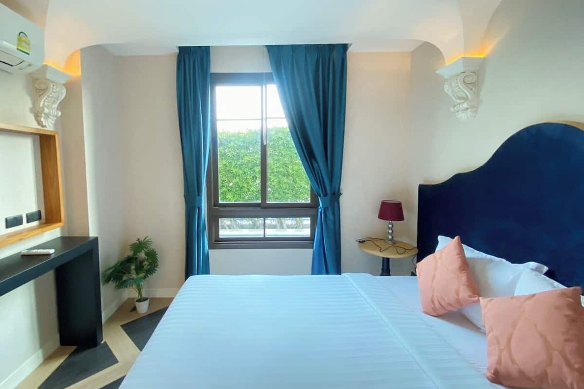 A hotel room with a large bed, a nightstand with a lamp, a window with curtains, and a view of greenery outside.