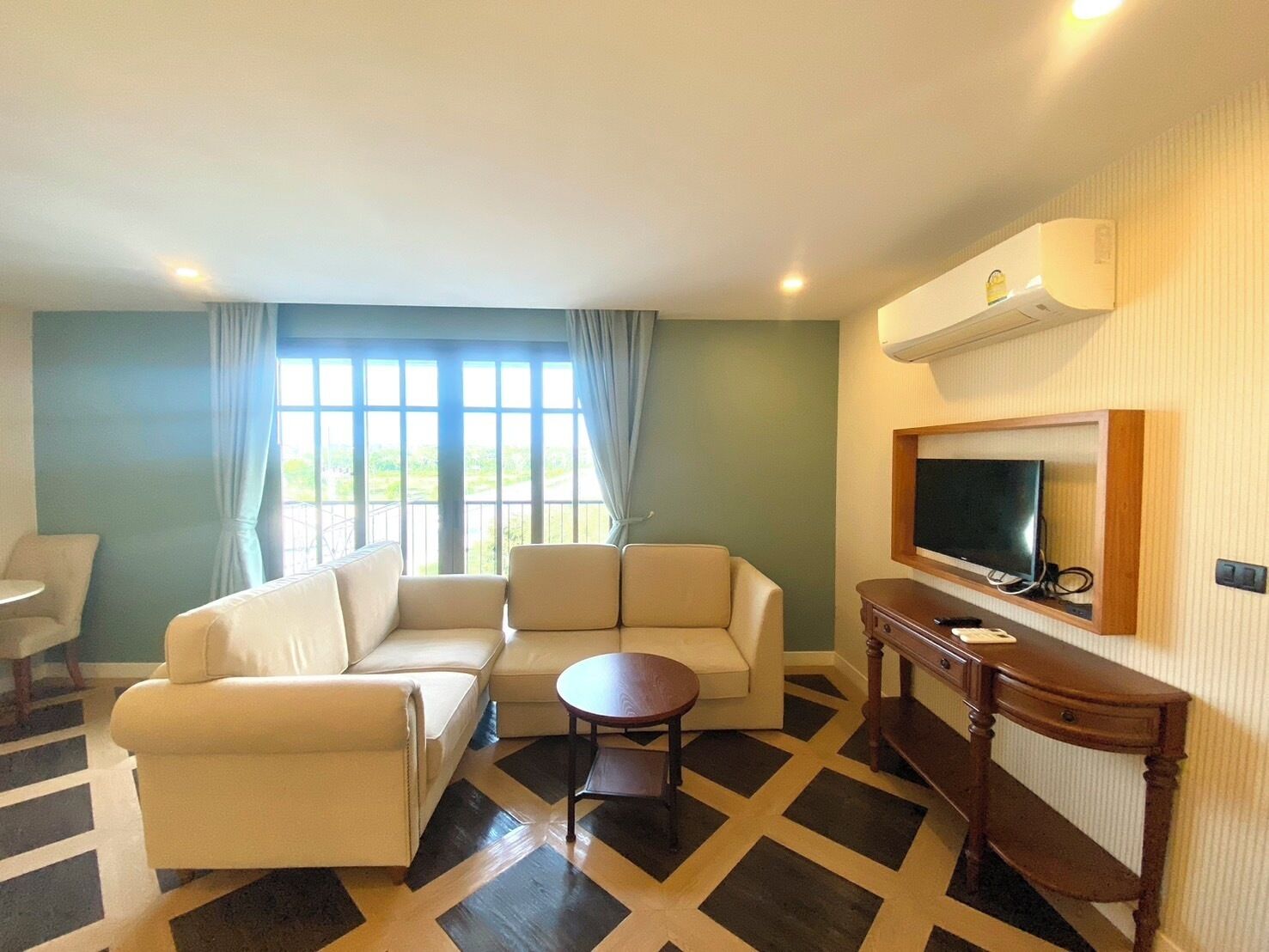 A hotel room with a sofa set, a small round table, a TV mounted on the wall, and a large window with curtains.