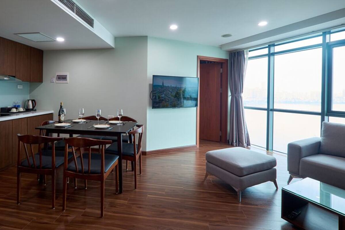 A modern kitchen with wooden cabinets, a dining table with chairs, a large window with a city view, and a grey sofa.