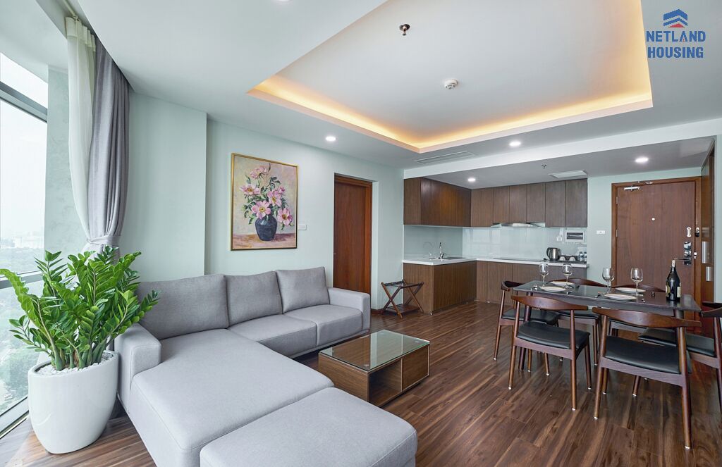 A modern living room with a grey sofa, a coffee table, and a dining area with wooden chairs and a glass table. There is a kitchen area with wooden cabinets and a sink.