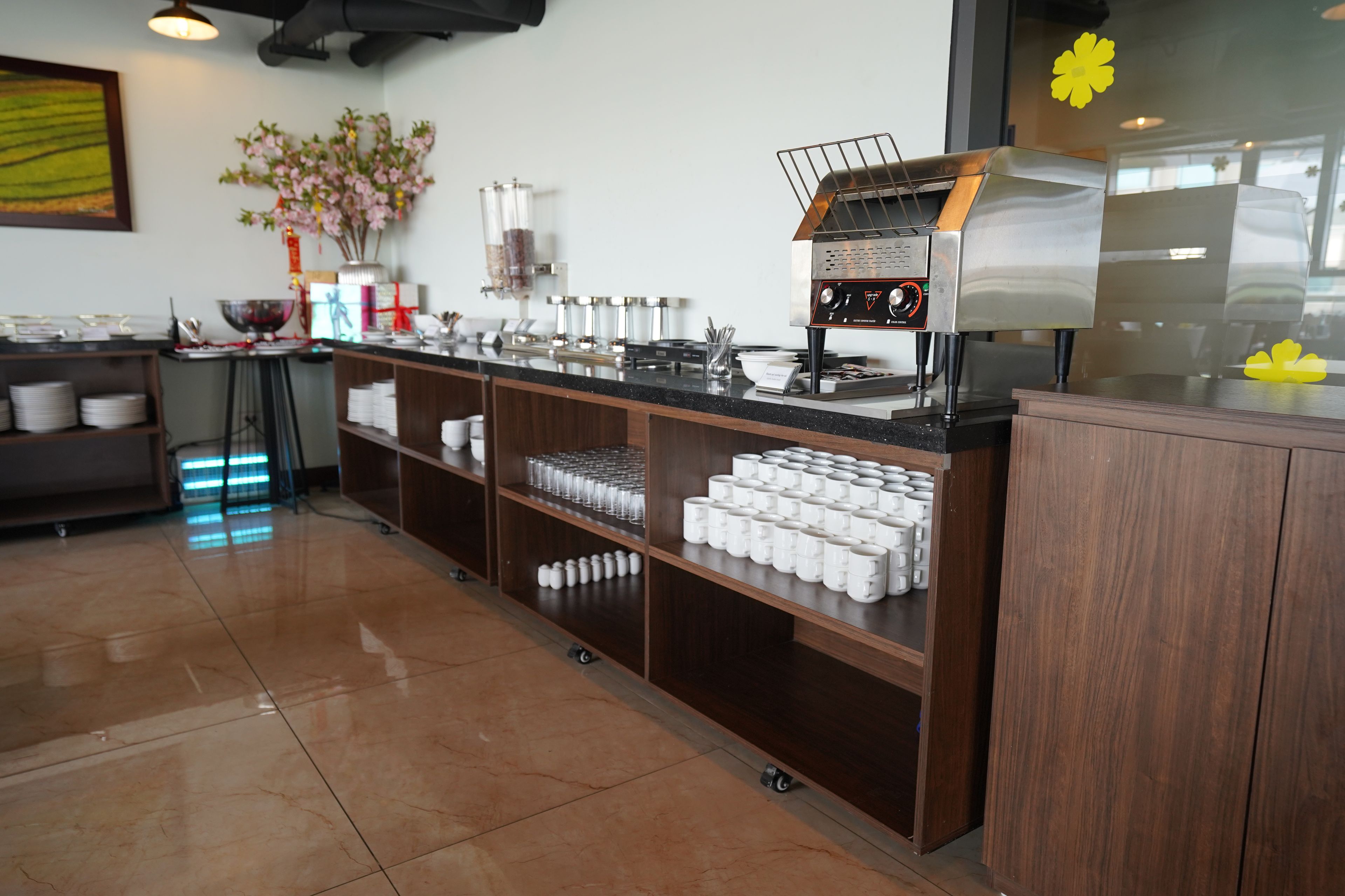 A clean, modern cafe with a coffee machine, shelves with cups, and a decorative plant.