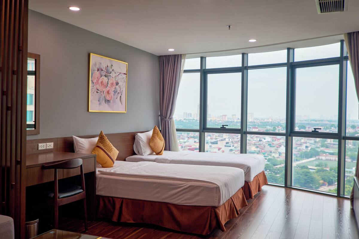 A hotel room with two beds, a desk, and a large window offering a city view.