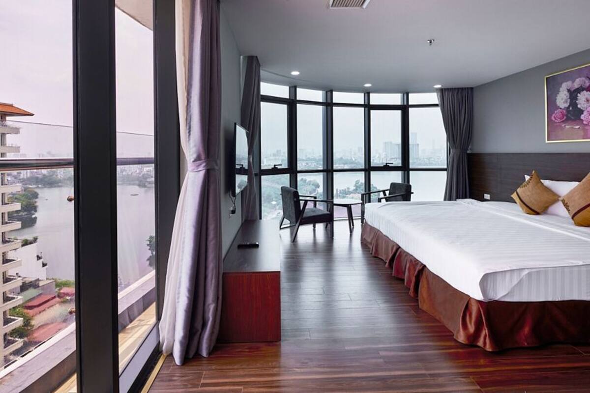 A hotel room with a large bed, a desk, and a chair. The room has a large window with a view of buildings and a body of water.