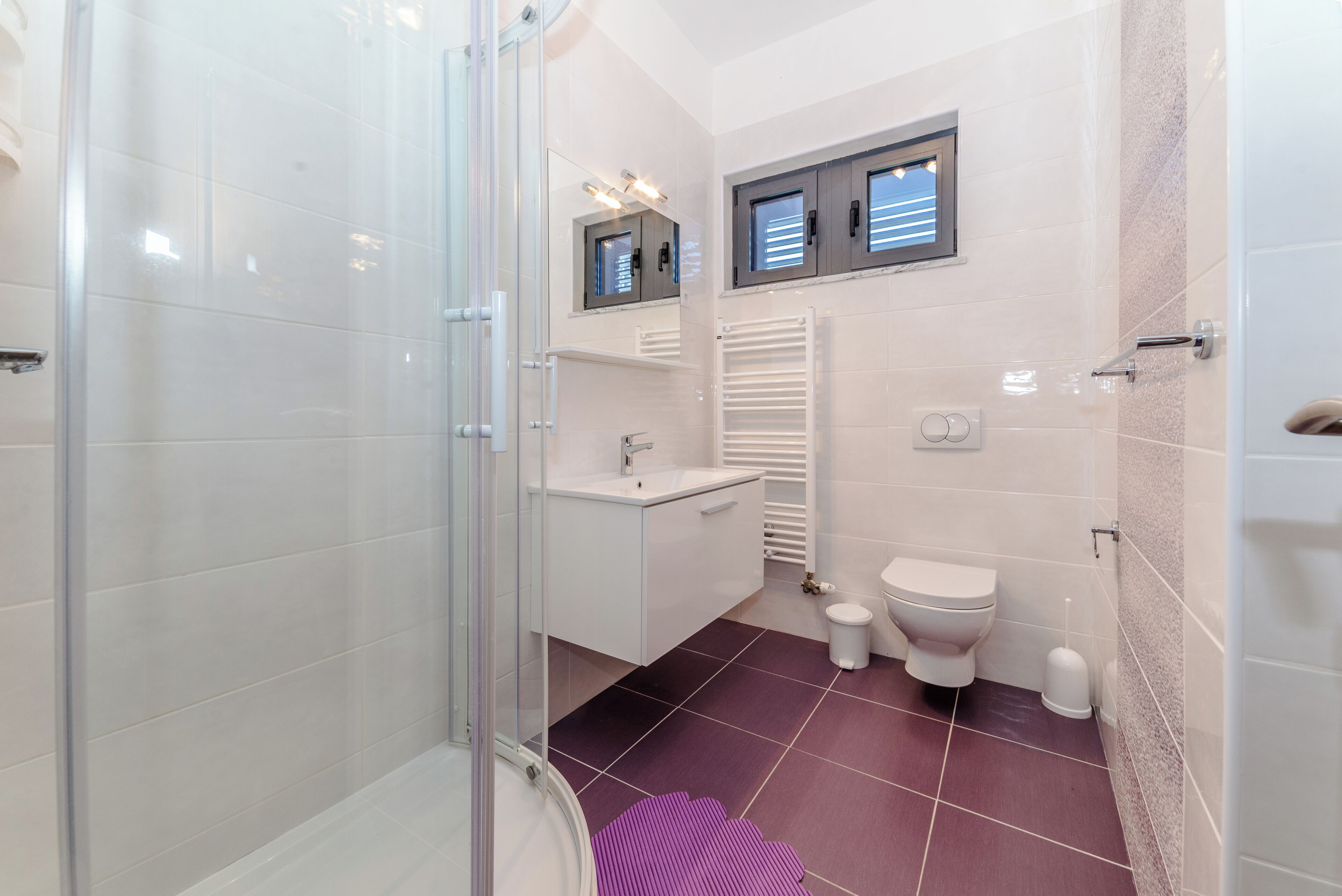 A modern bathroom with a glass shower enclosure, a white sink, a toilet, and a window above the toilet.