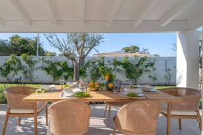 An outdoor dining area with wicker chairs and a wooden table set for a meal, surrounded by a white wall with greenery. - Charming Villa in Fontane Bianche, Italy - Perfect for Families (Syracuse)