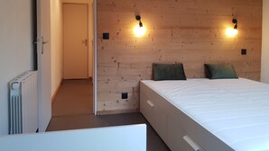 Een slaapkamer met een houten hoofdeinde, twee bedlampen, een bed met wit beddengoed en een deur naar een andere kamer.