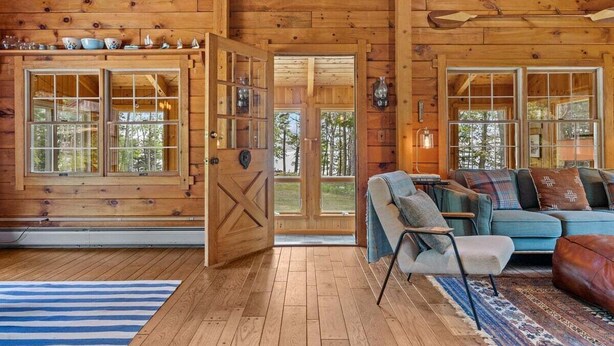 Interior de una cabaña de madera con piso de madera, un sofá, una silla y una mesa de comedor.