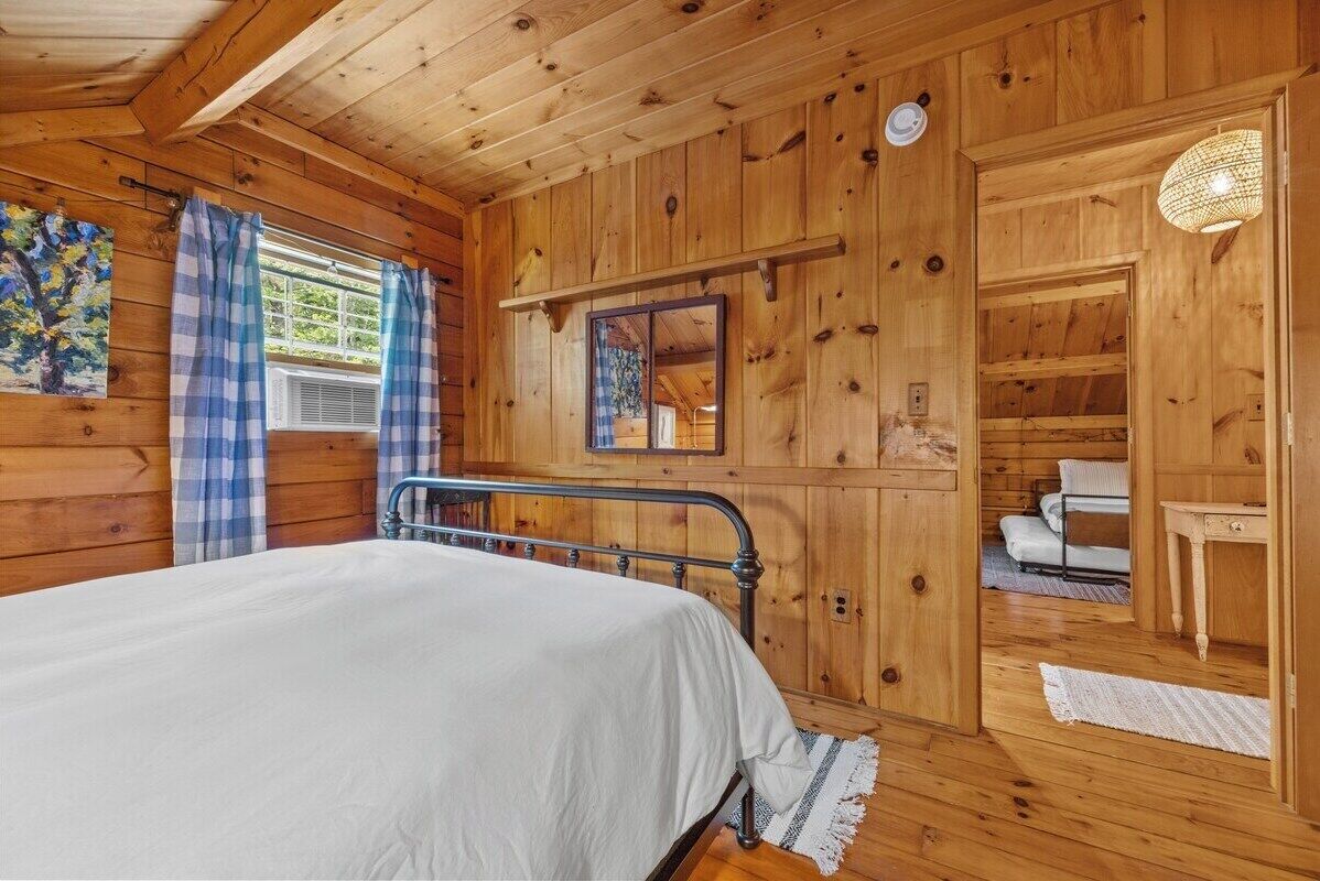 Quarto de cabana de madeira com cama, janela com cortinas, espelho e uma mesinha.