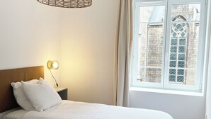 A bedroom with two beds, a hanging light fixture, a nightstand with a lamp, and a window with a view of a building.