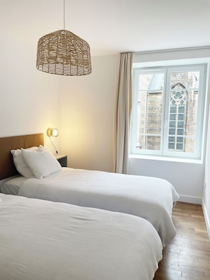 Een slaapkamer met twee bedden, een hanglamp, een nachtkastje met een lamp en een raam met uitzicht op een gebouw.