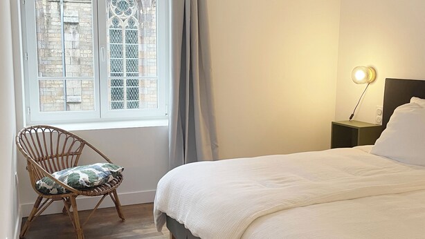 A bedroom with a bed, a chair, a window with curtains, a hanging light fixture, and a nightstand with a lamp.