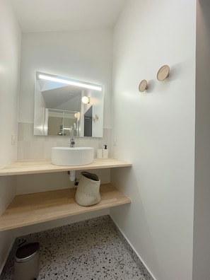 Een badkamer met een wit wastafel, een spiegel en een lamp erboven. Twee ronde decoraties hangen aan de muur en er staat een prullenbak.