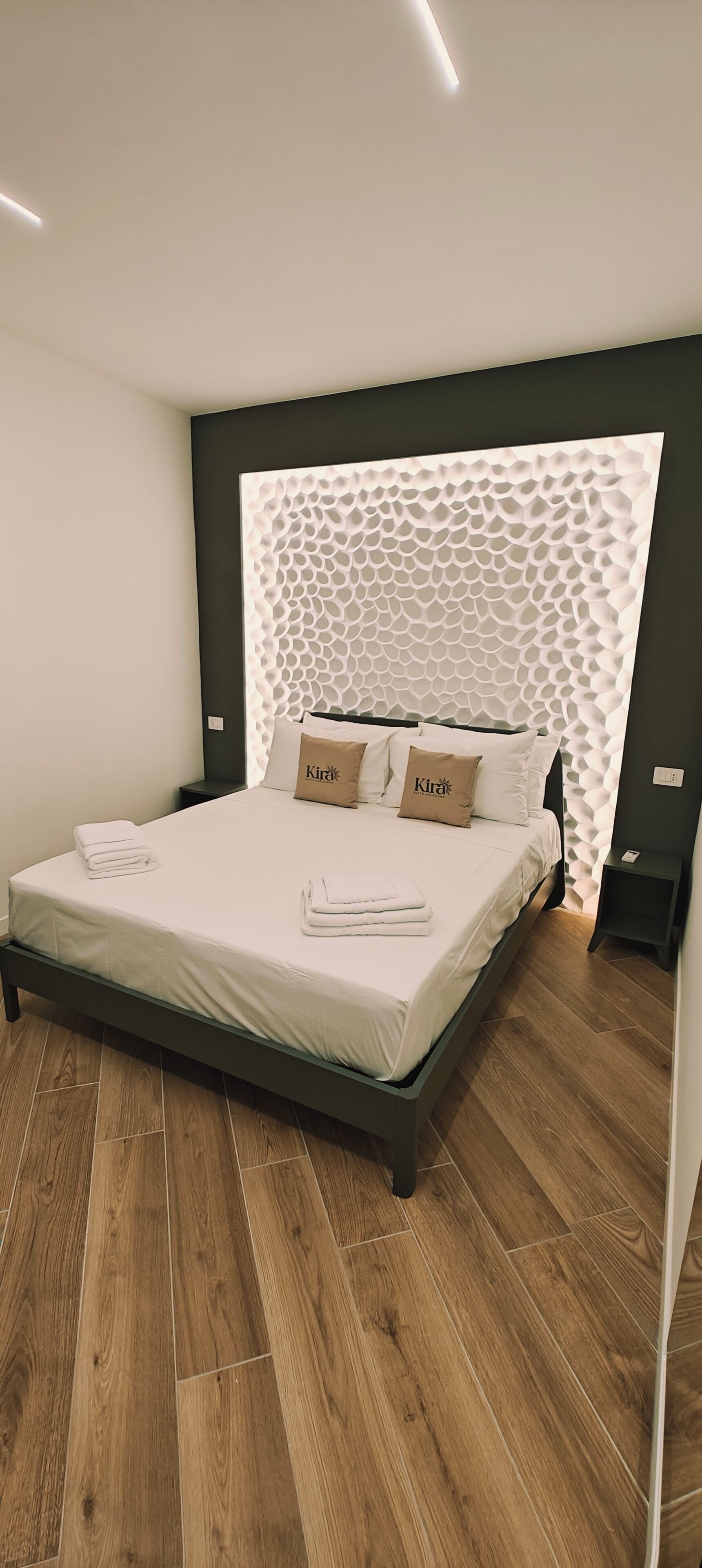 Une chambre moderne avec un plancher en bois, un lit avec une literie blanche et deux oreillers, une tête de lit sombre et une table de chevet.