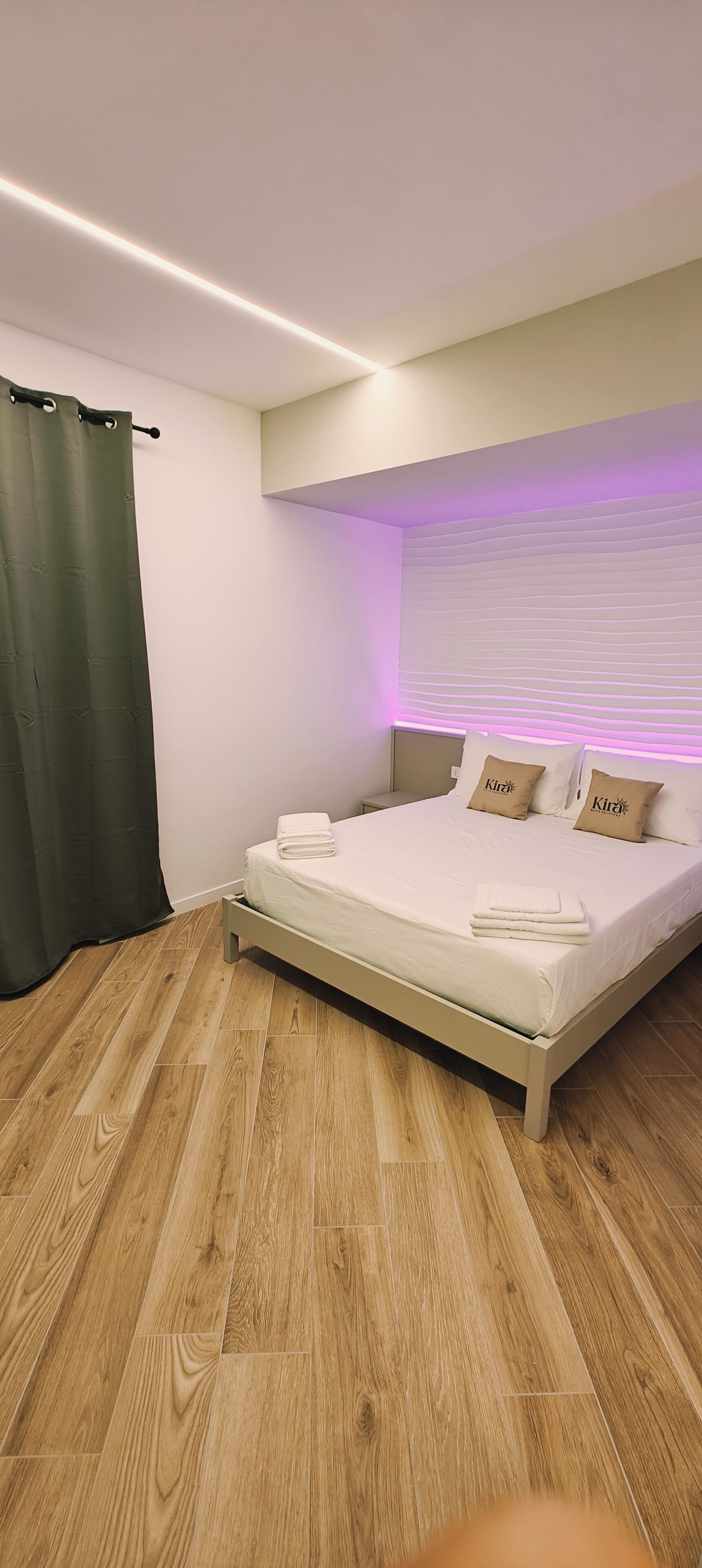 A modern bedroom with a wooden floor, a bed with white bedding and two pillows, a nightstand, and a window with a curtain.
