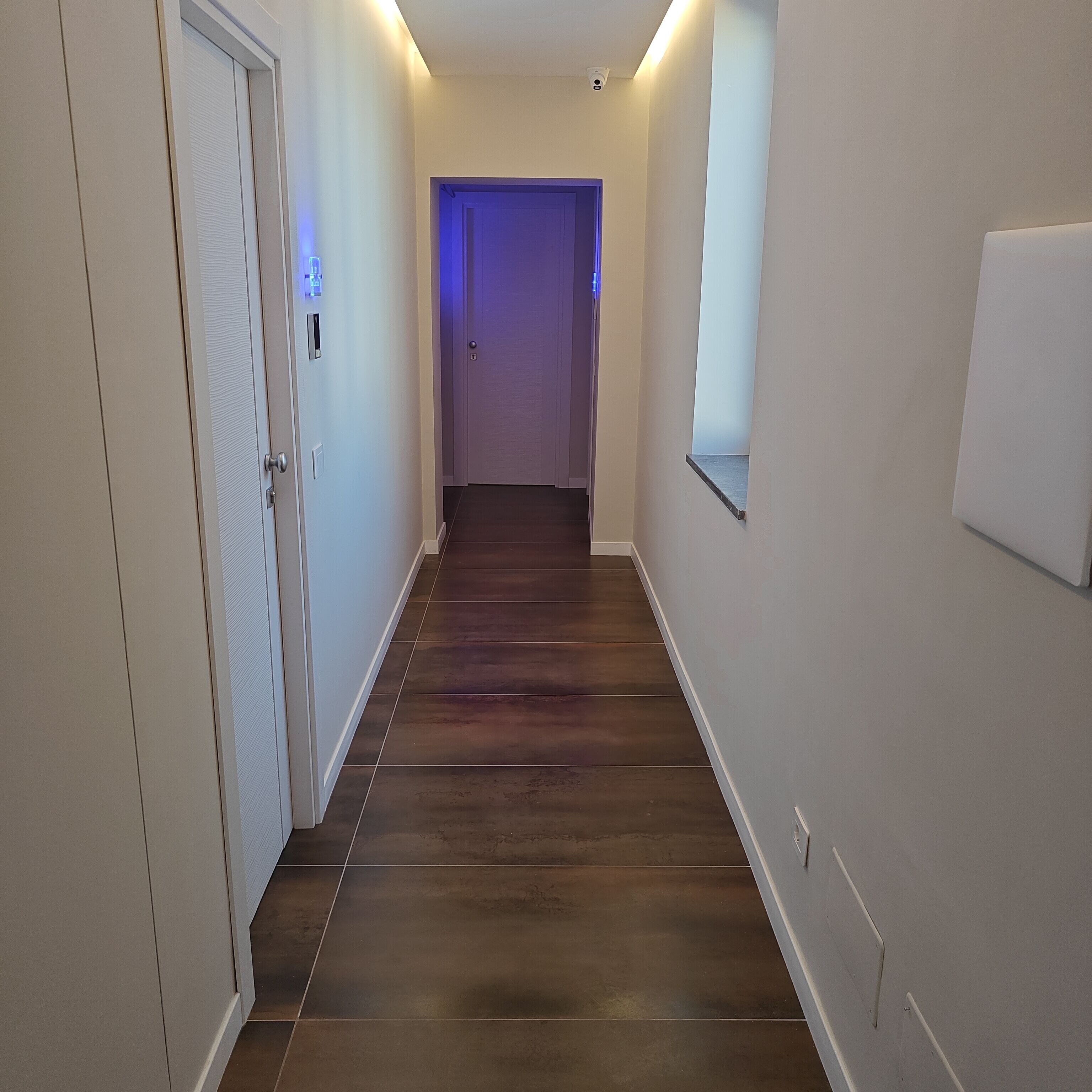 A hallway with wooden flooring, white walls, and a door with a light switch.