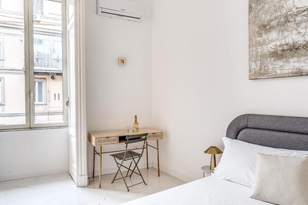 Habitación de hotel con cama, escritorio con una botella y vasos, lámpara y un cuadro en la pared.