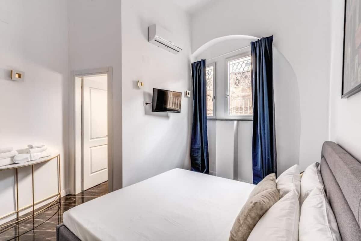 A hotel room with a bed, a TV, a window with curtains, a door, and a small table with towels.