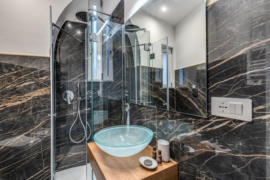 Une salle de bain moderne dotée d’une douche à parois de verre, d’un mur recouvert de carreaux de marbre et d’un meuble-lavabo en bois avec un vasque en verre.
