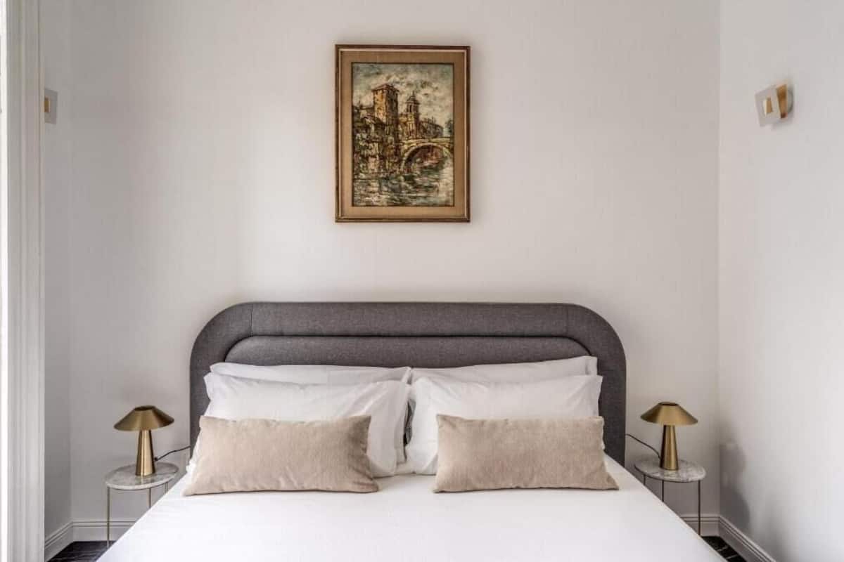 A neatly made bed with a grey headboard, two bedside lamps, and a framed cityscape painting on the wall.