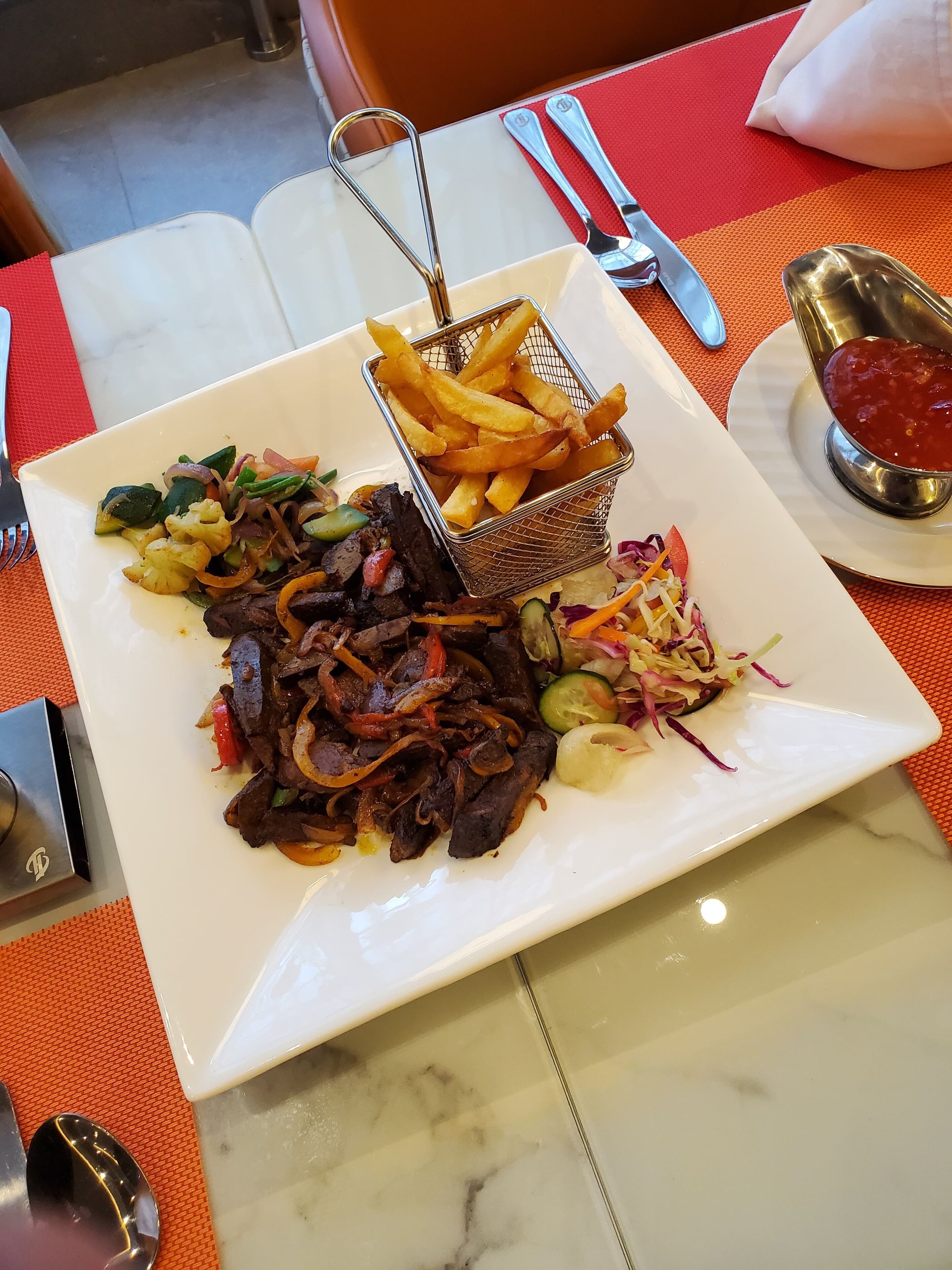 A dining table with a white plate of food, a basket of fries, and a small bowl of sauce.