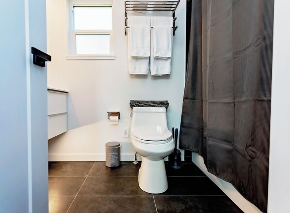A modern bathroom with a toilet, a trash bin, a towel rack with towels, a window, and a shower curtain. - Bumbleberry 3 Bedroom Guest Home
New Executive home in the heart of Kitimat BC (Kitimat)