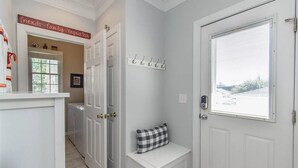 Un couloir avec une porte blanche, un banc doté d’un coussin à carreaux, un porte-manteau et une pancarte au-dessus de la porte.