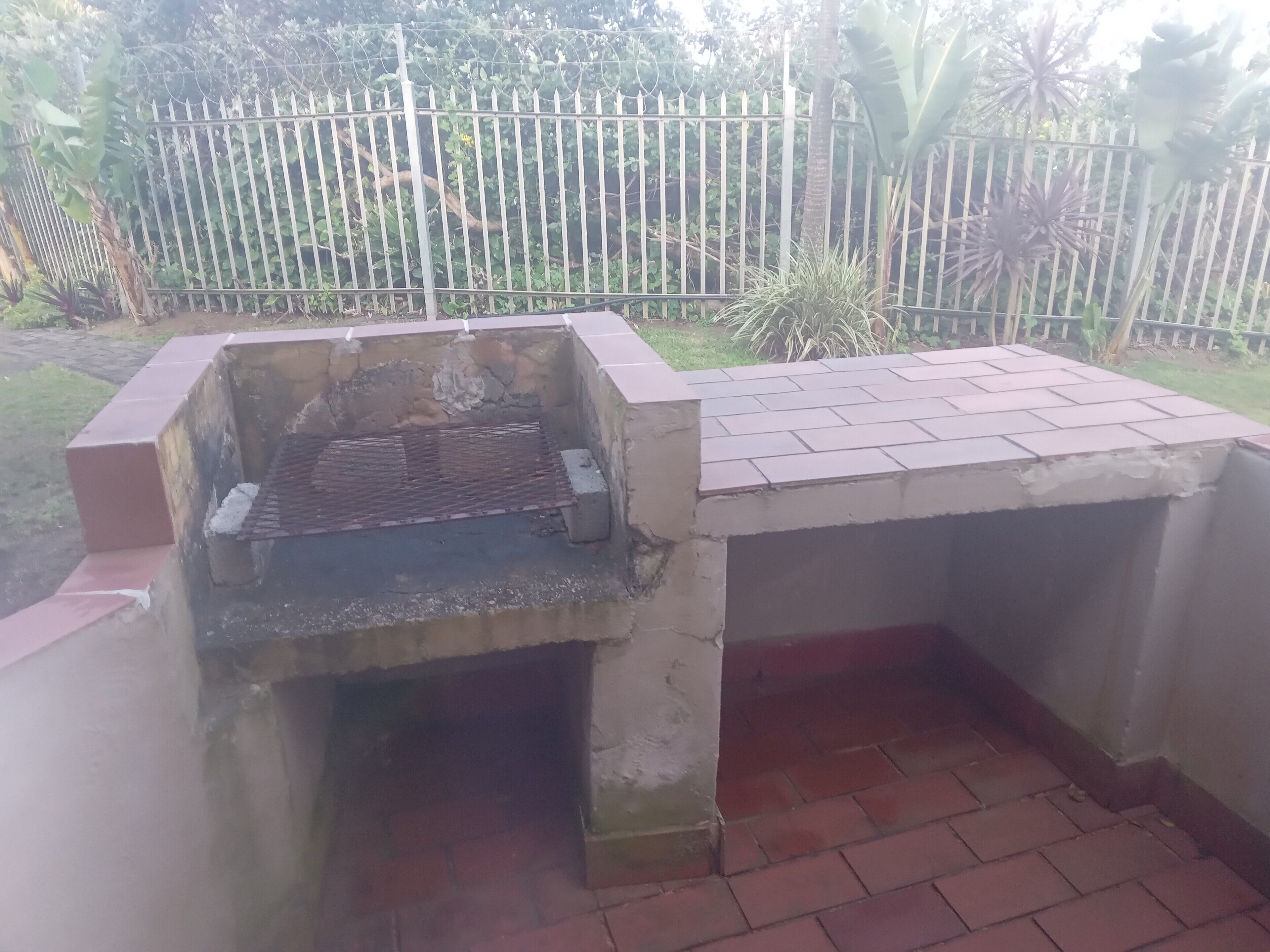 Un espace barbecue extérieur en béton, doté d’une grille en métal, entouré d’un sol en briques et d’une clôture en métal.