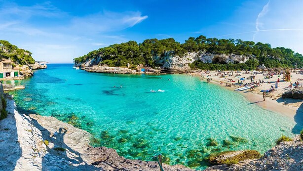 Een kustgezicht met helder turquoise water, rotskliffen en een strand met mensen en boten.