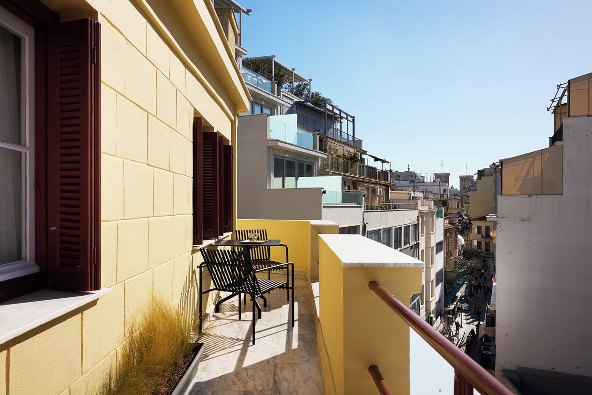 A narrow urban street with buildings on either side, a balcony with a chair and table, and a clear blue sky.