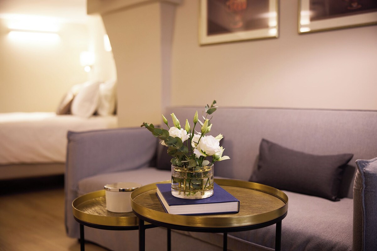 Une pièce comprenant un lit, un canapé, une petite table ronde avec un livre et un vase de fleurs, ainsi qu’un mur orné de tableaux encadrés.