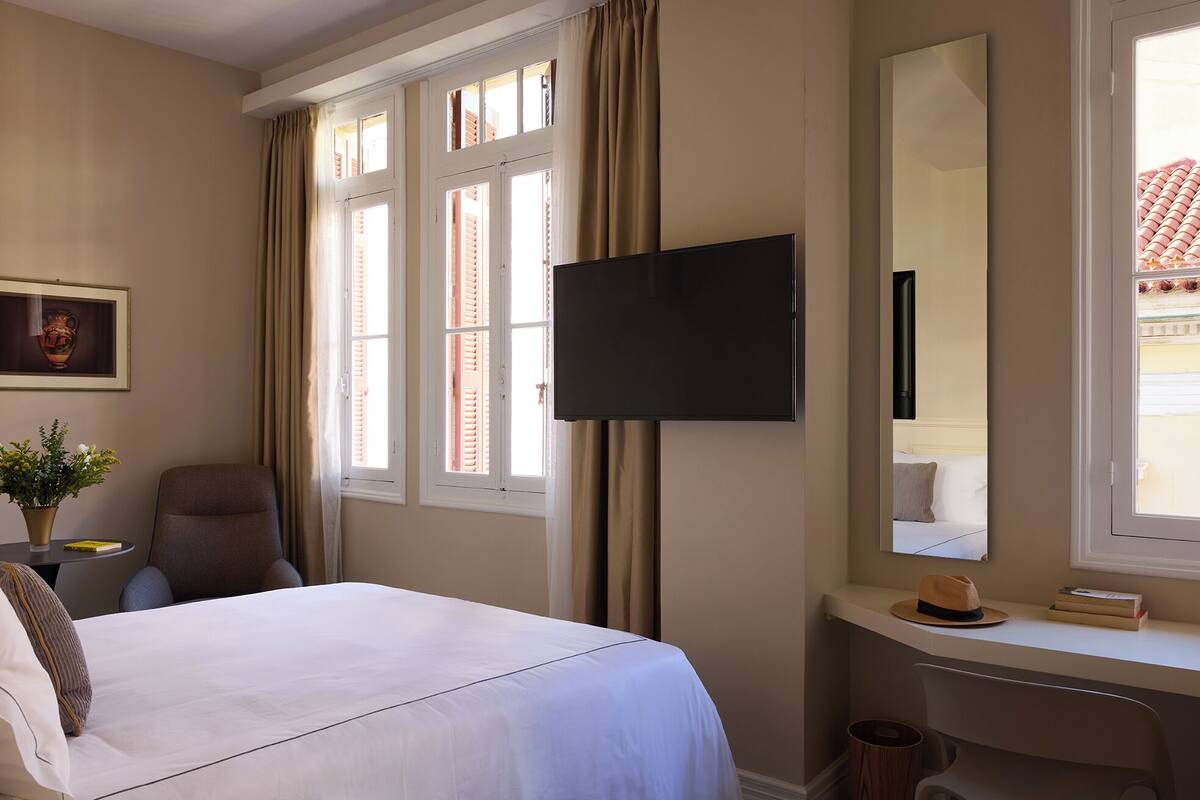 Ett hotellrum med en säng, ett skrivbord, en stol, en tv och ett fönster med gardiner.