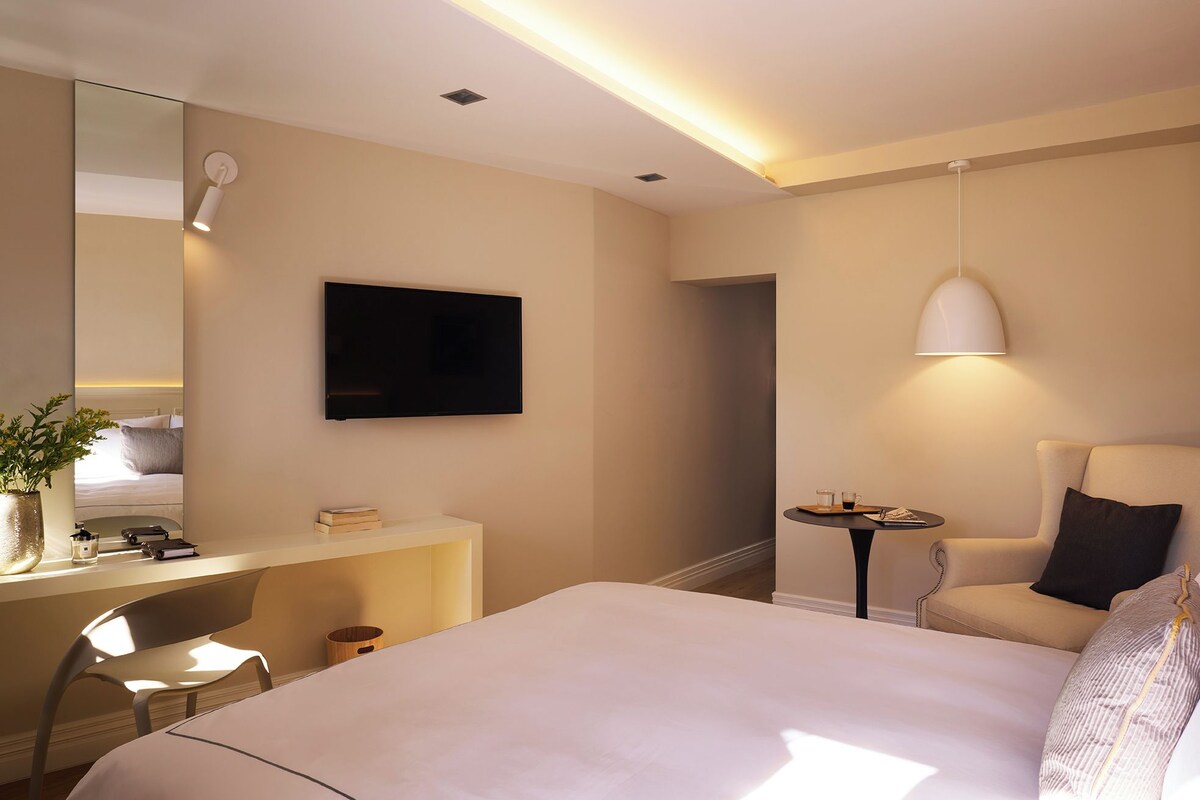 A hotel room with a bed, a desk with a chair, a TV, a lamp, and a small table with a chair.