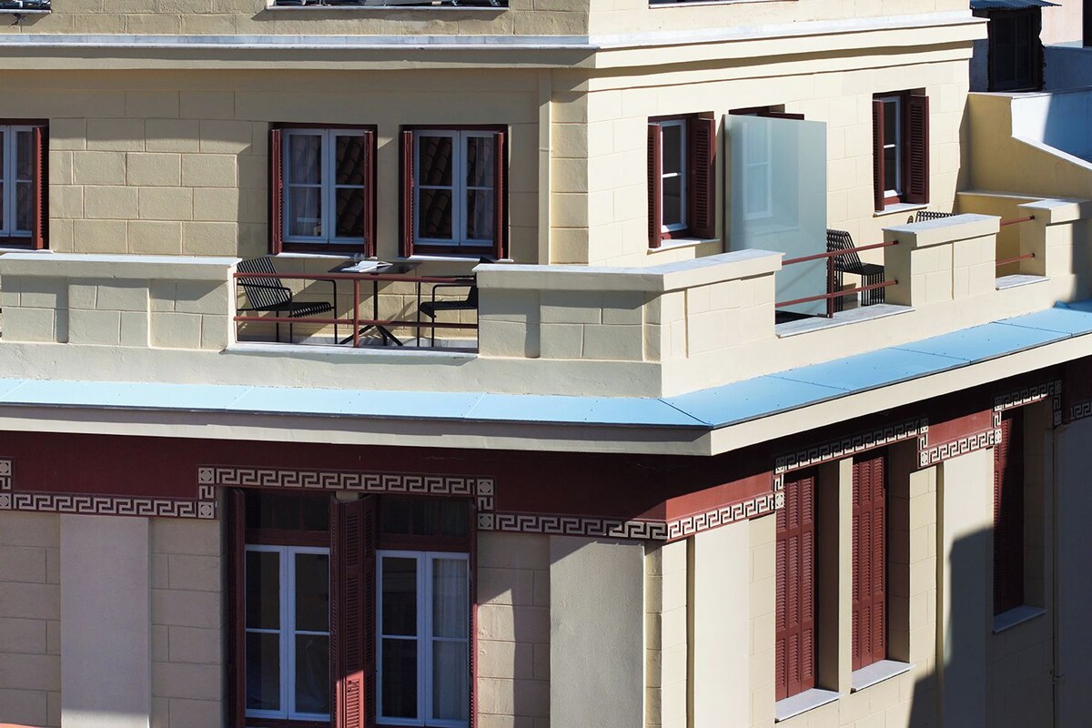 A multi-story building with a flat roof terrace, multiple windows with shutters, and a balcony with outdoor furniture.