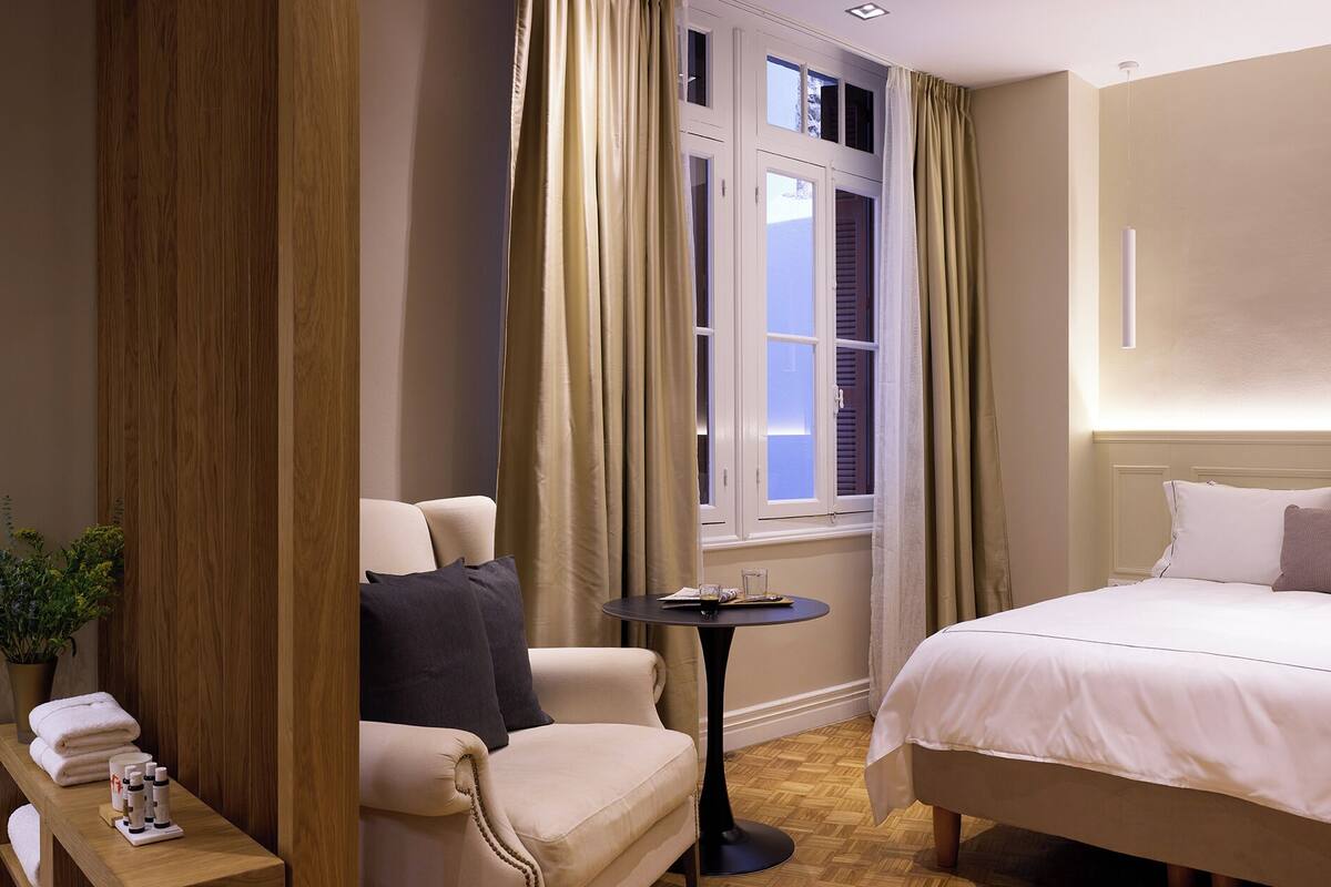 Ett hotellrum med en säng, en stol, ett litet bord och ett fönster med gardiner.