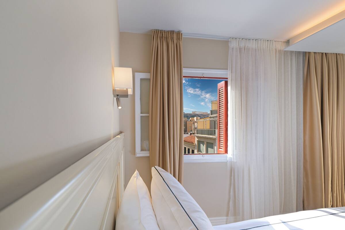 Ett hotellrum med en säng, ett fönster med gardiner, en vägglampa och utsikt över byggnader utanför.