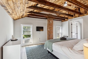 Une chambre avec un plafond en bois, une colonne en briques, un lit recouvert d’une couverture verte et un téléviseur à écran plat fixé au mur.