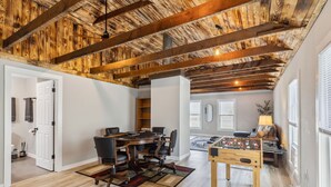 Amplia habitación con techo alto que presenta vigas de madera a la vista, zona de comedor con mesa redonda y sillas, y una mesa de fútbol de mesa.